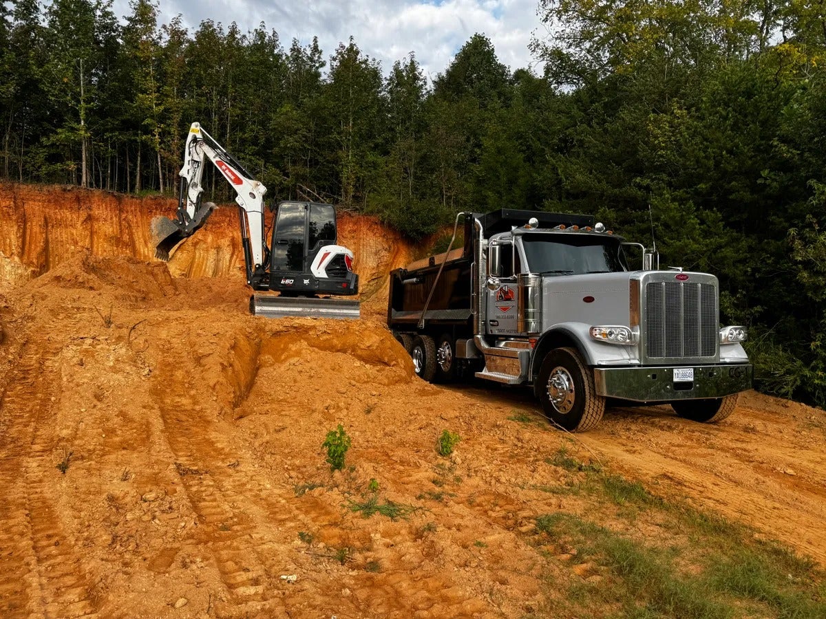 An excavator digging into an orange dirt hill next to a silver dump truck, with a forest in the background.