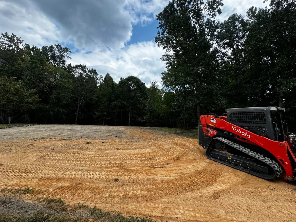 A red Kubota skid steer loader parked on a newly graded, mostly flat, sandy lot with a dark green forest backdrop.