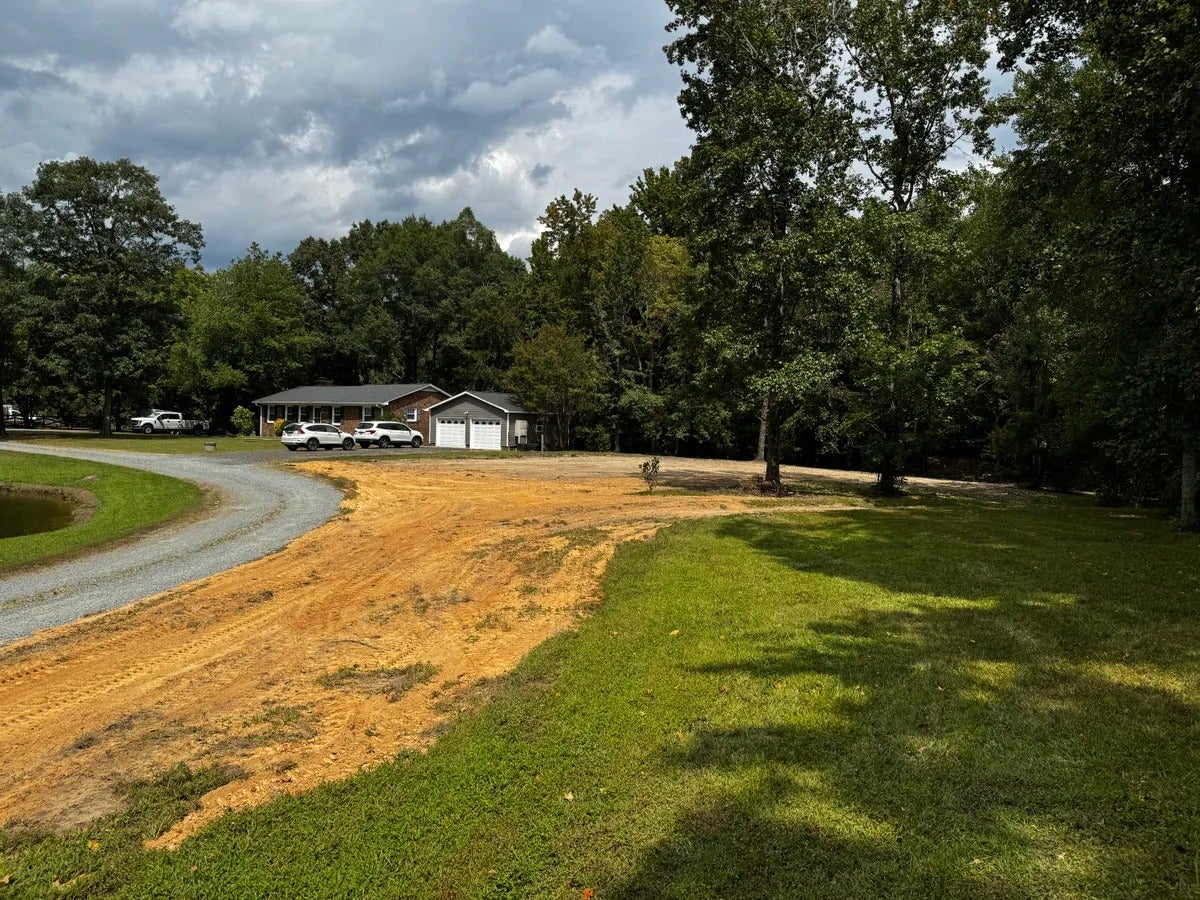 Rural house with attached garage surrounded by green trees, a dirt patch, and a gravel driveway.