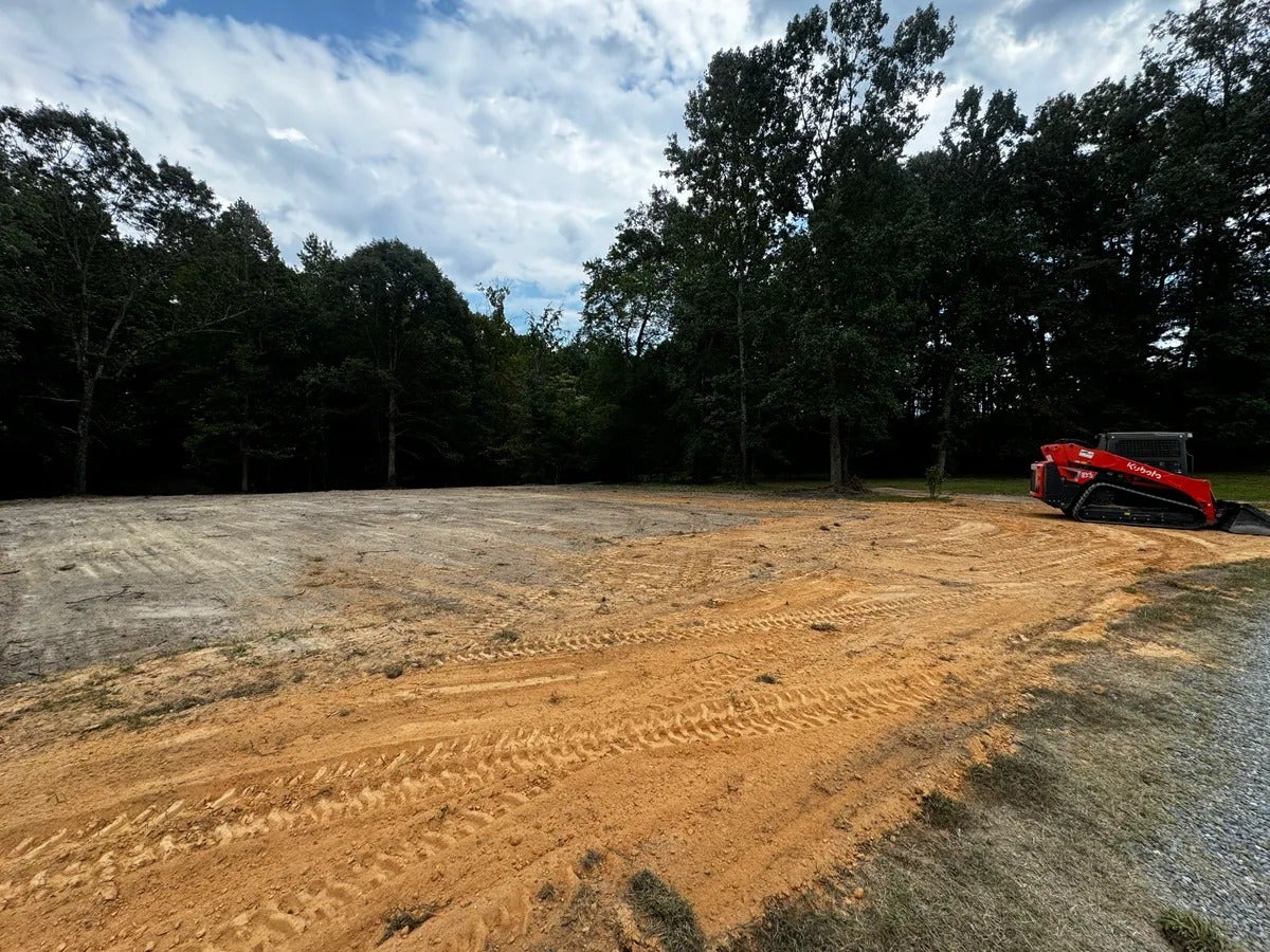 A red Kubota skid-steer loader sits on a dirt lot with tire tracks, surrounded by trees under a cloudy sky.