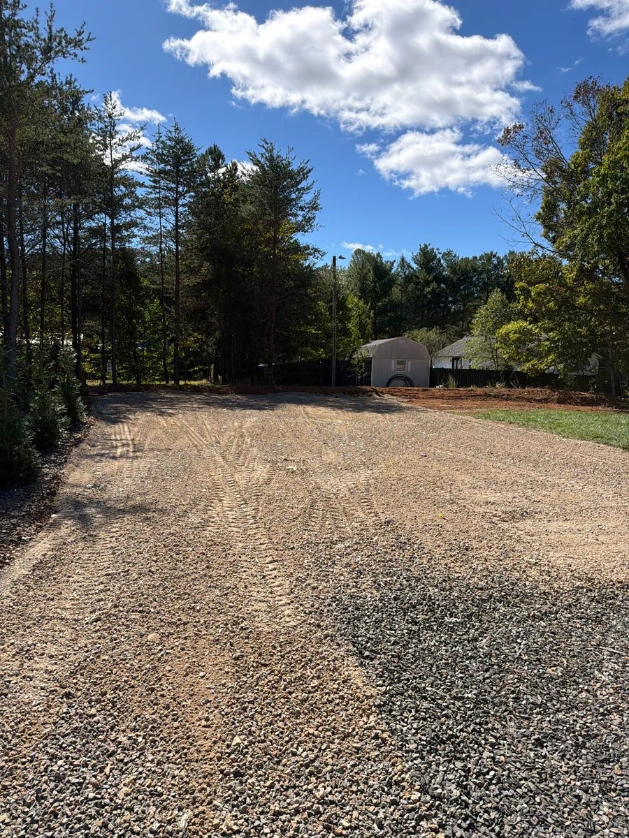 A new gravel driveway and parking area with fresh tire tracks, bordered by trees, leading to a small shed.