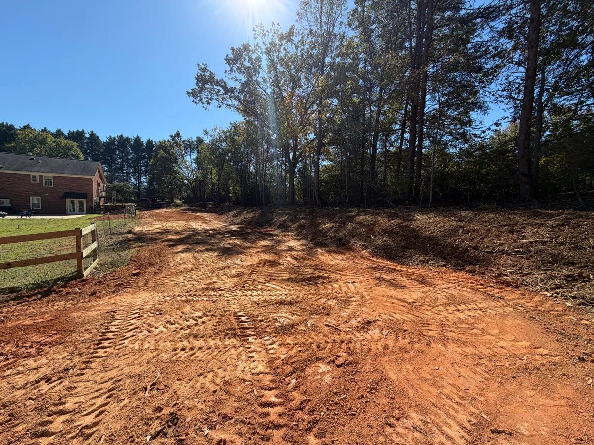 Excavated red dirt area with tire tracks, bordered by a wooden fence, house, and dense woods under a bright sky.