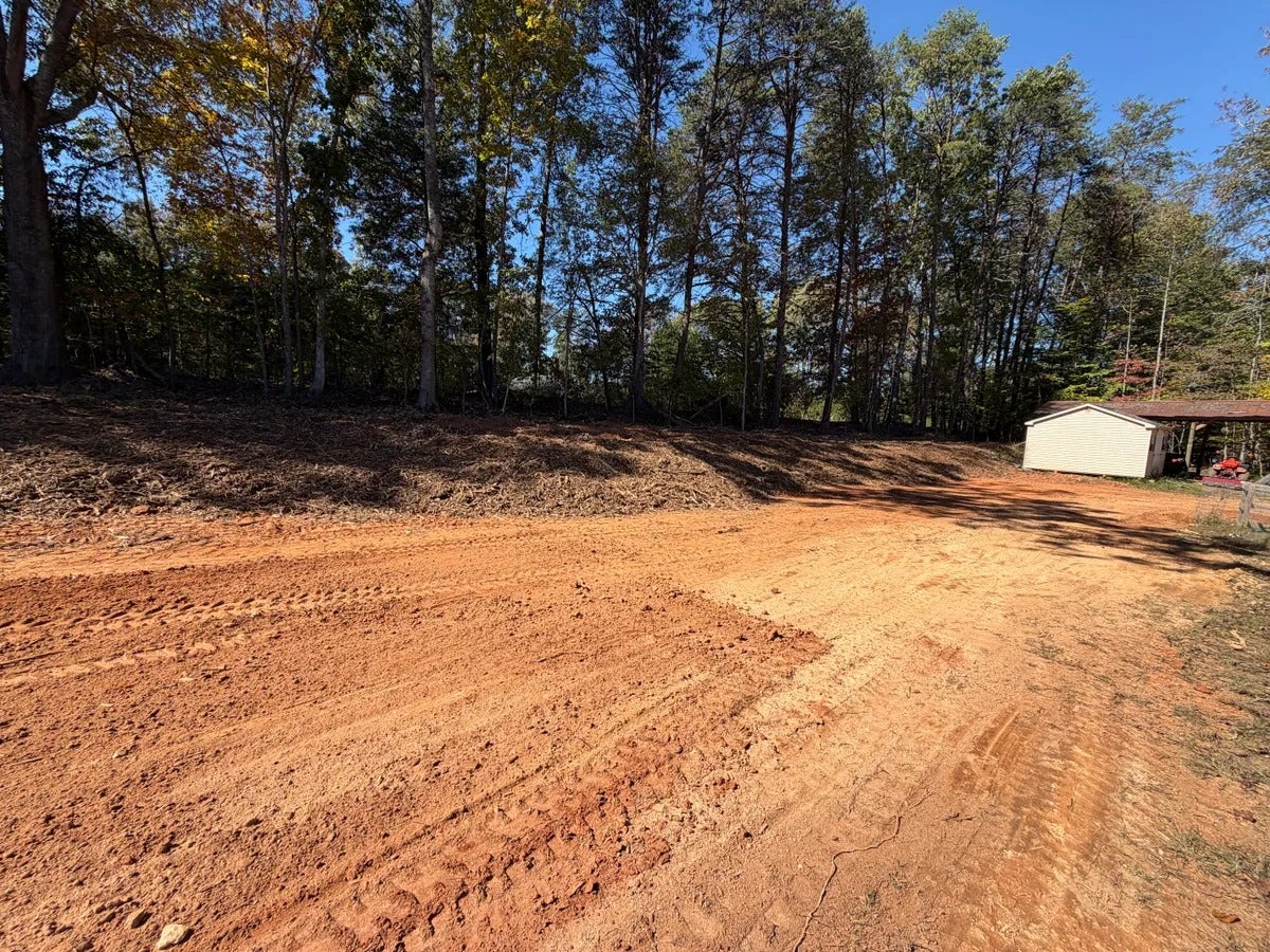 Cleared, orange-red dirt lot with tire tracks, backed by a wooded hill and a small white shed.