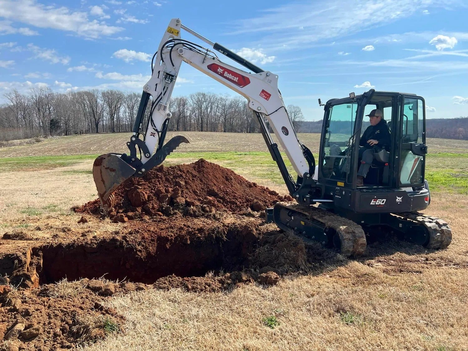 Man operating Bobcat E60 excavator digging a trench in dry grass