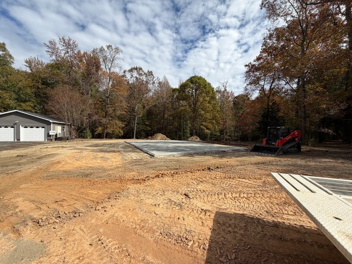 Cleared land with a concrete foundation, a house with a two-car garage, and an excavator among autumn trees.