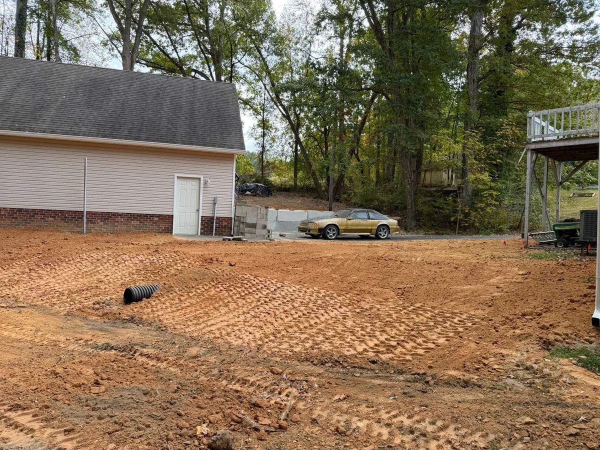 A side view of a beige garage with a gold Ford Mustang parked nearby, set against a backdrop of trees.
