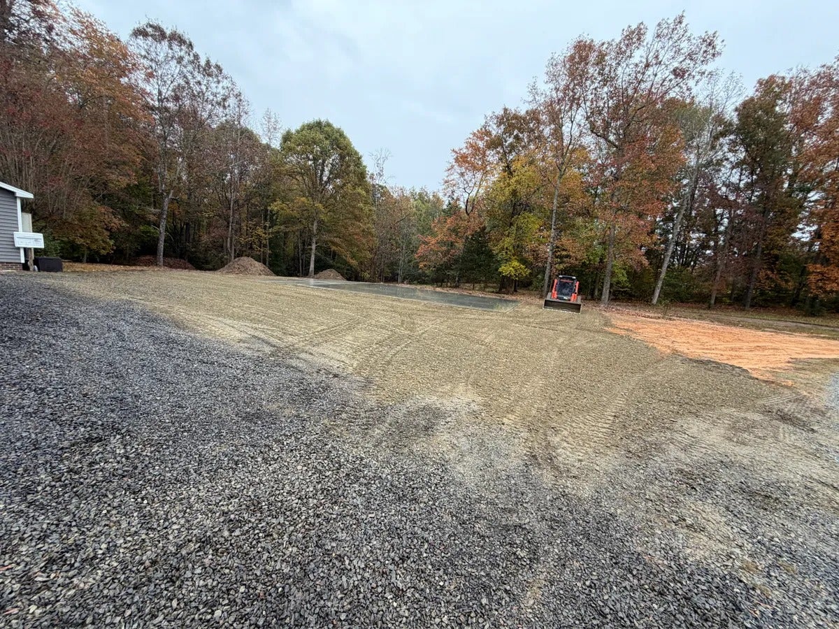 A large area of dark gray gravel transitioning to lighter-colored gravel and a small section of dirt, with trees and an orange skid steer in the background.