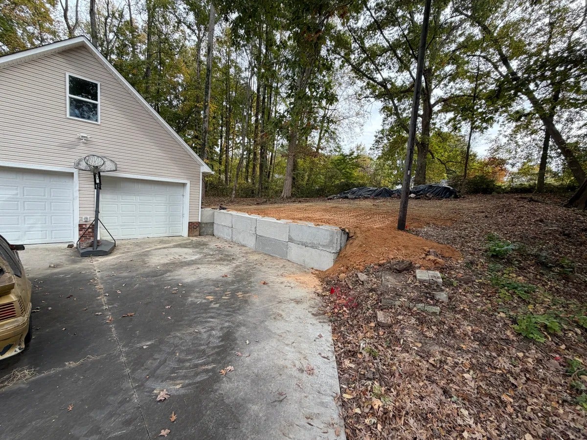 A driveway beside a two-car garage with a basketball hoop, retaining wall, and excavated dirt.