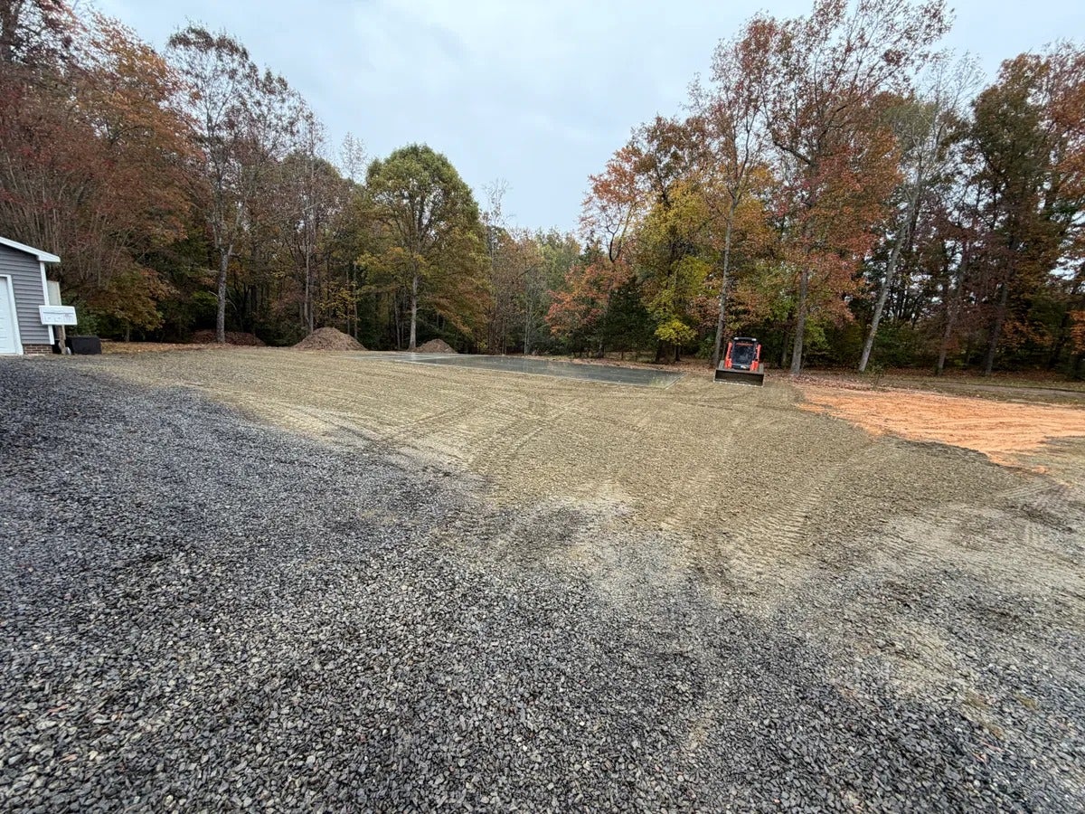 Skid-steer loader grading a gravel and dirt lot, with autumn trees and a shed in the background.