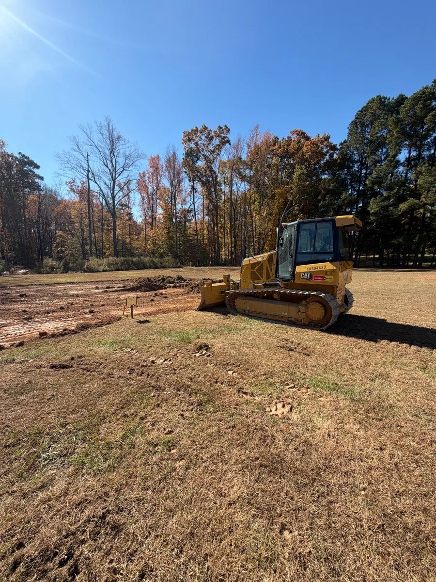 Yellow bulldozer parked on dried grass next to a mound of dirt with trees in the background under a blue sky.