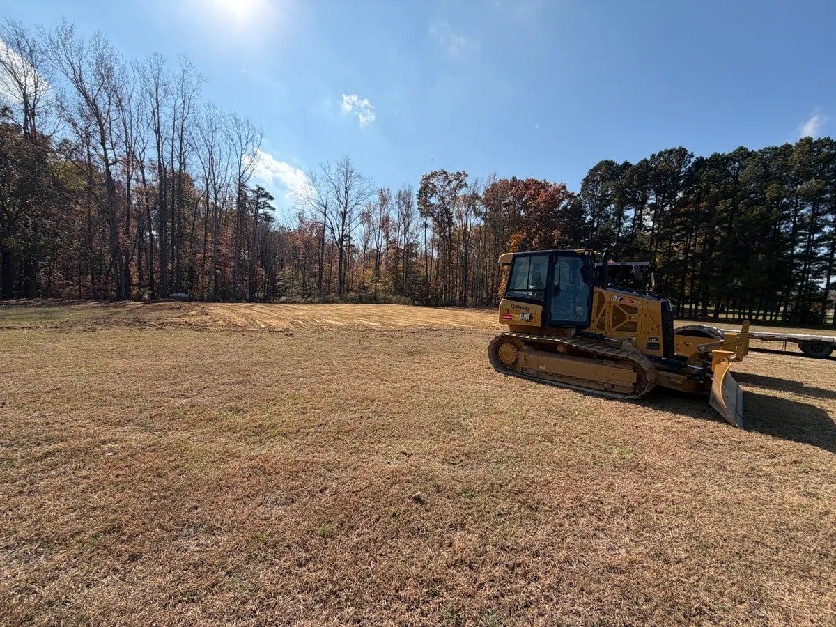 A yellow bulldozer sits on a field of dry grass, near a treeline under a bright blue sky.