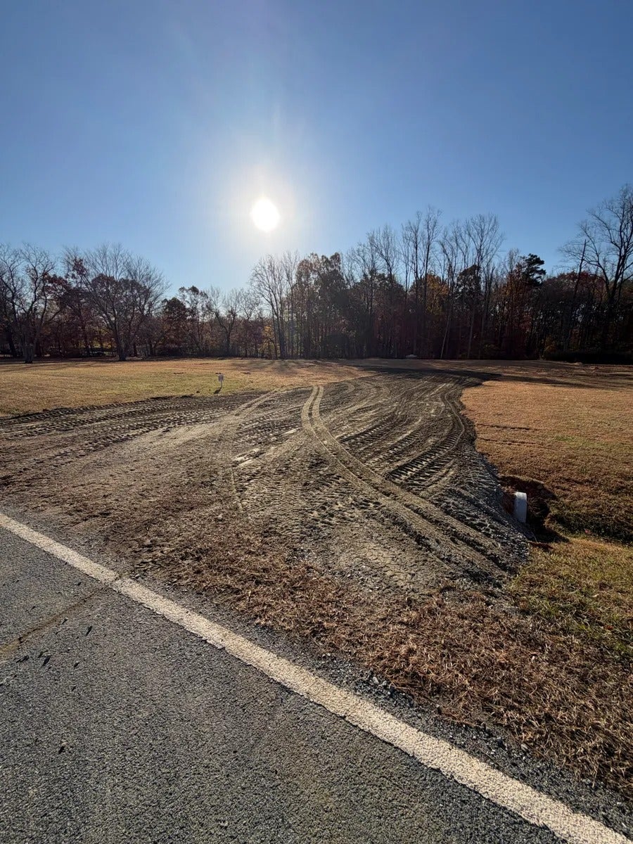 An asphalt road with a white line next to a dirt construction entrance with tire tracks, leading to a field and forest.