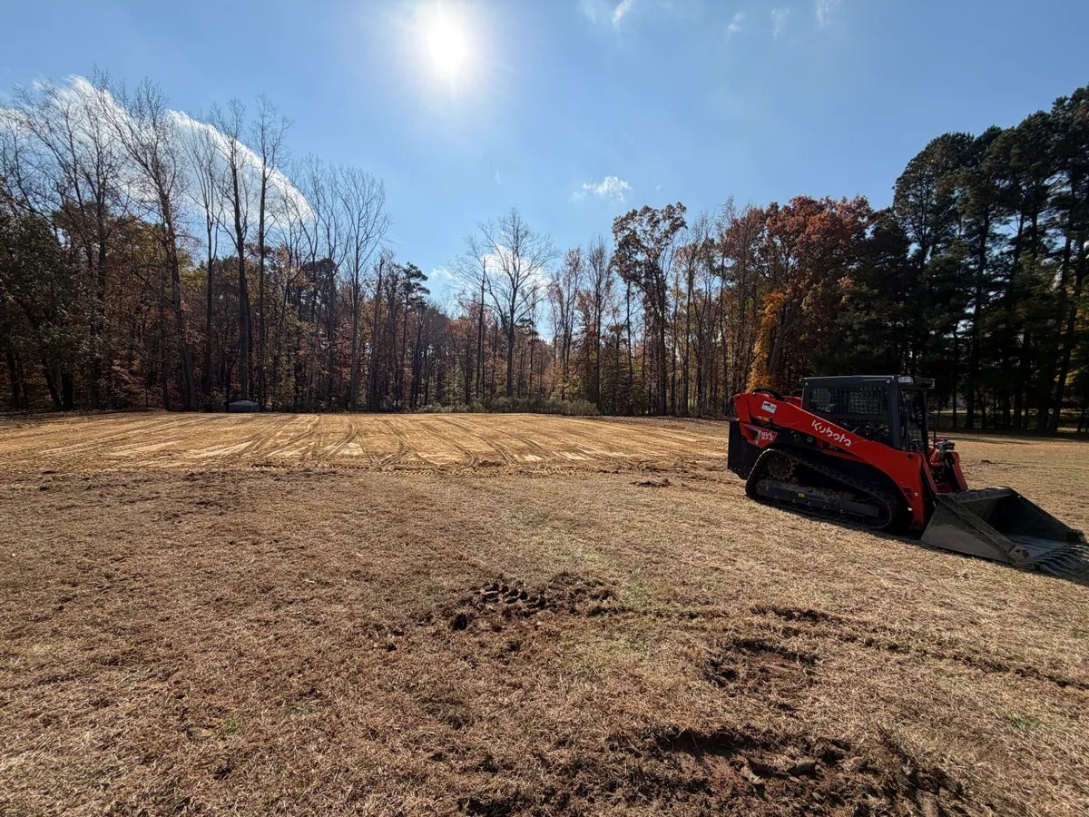 Red Kubota skid-steer with a bucket attachment, parked on a cleared, uneven brown field under a sunny sky.