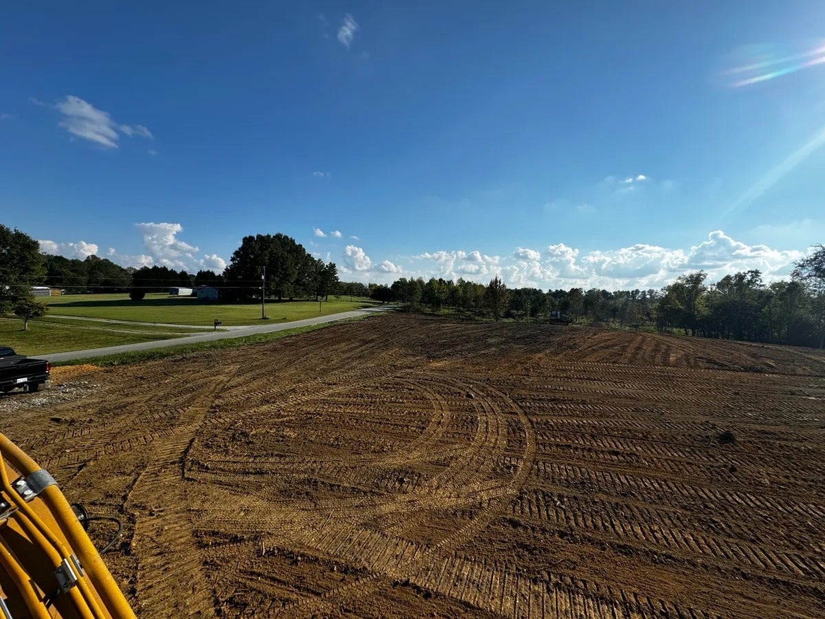 Construction site with freshly graded brown earth covered in tire tracks, under a blue sky with white clouds.