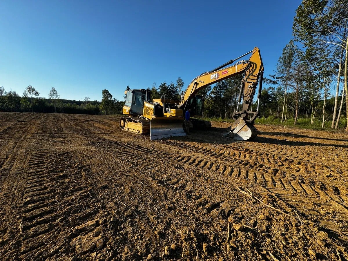 A yellow CAT excavator with tracks on cleared brown earth under a bright blue sky, with trees in the distance.