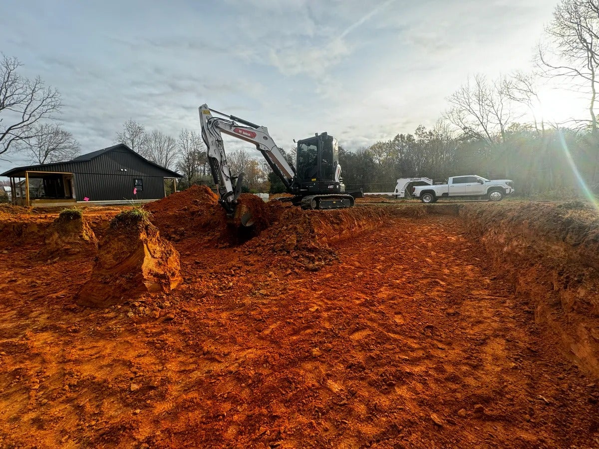 An excavator digs a foundation pit in red clay next to a dark barn and a white pickup truck.