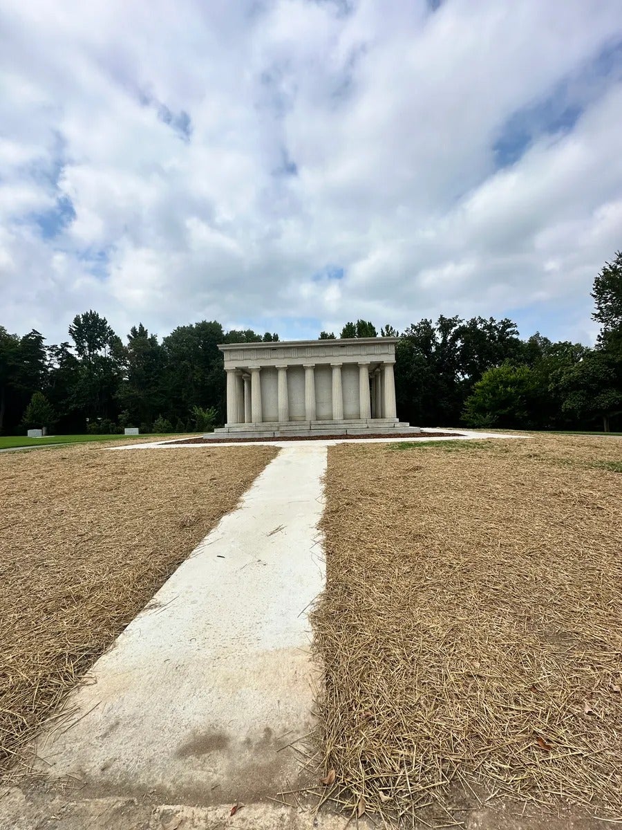 A paved path leads to a white, classical-style stone building with columns, surrounded by brown straw and green trees.