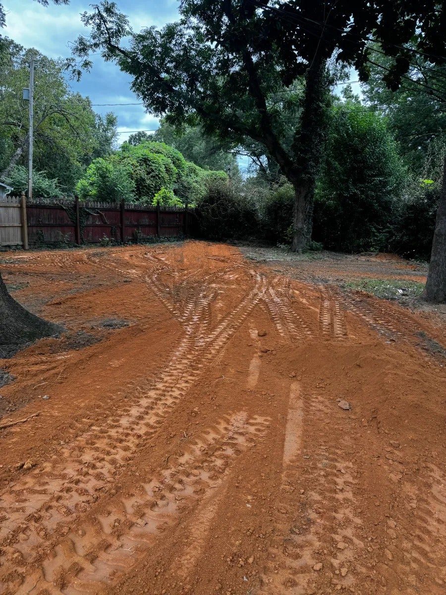 Reddish-brown dirt lot with tire tracks, surrounded by a wooden fence and lush green trees under a cloudy sky.