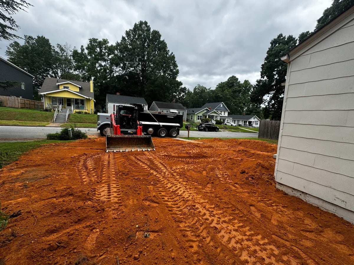 A construction site with disturbed orange dirt, a red skid-steer loader, and a large dump truck.