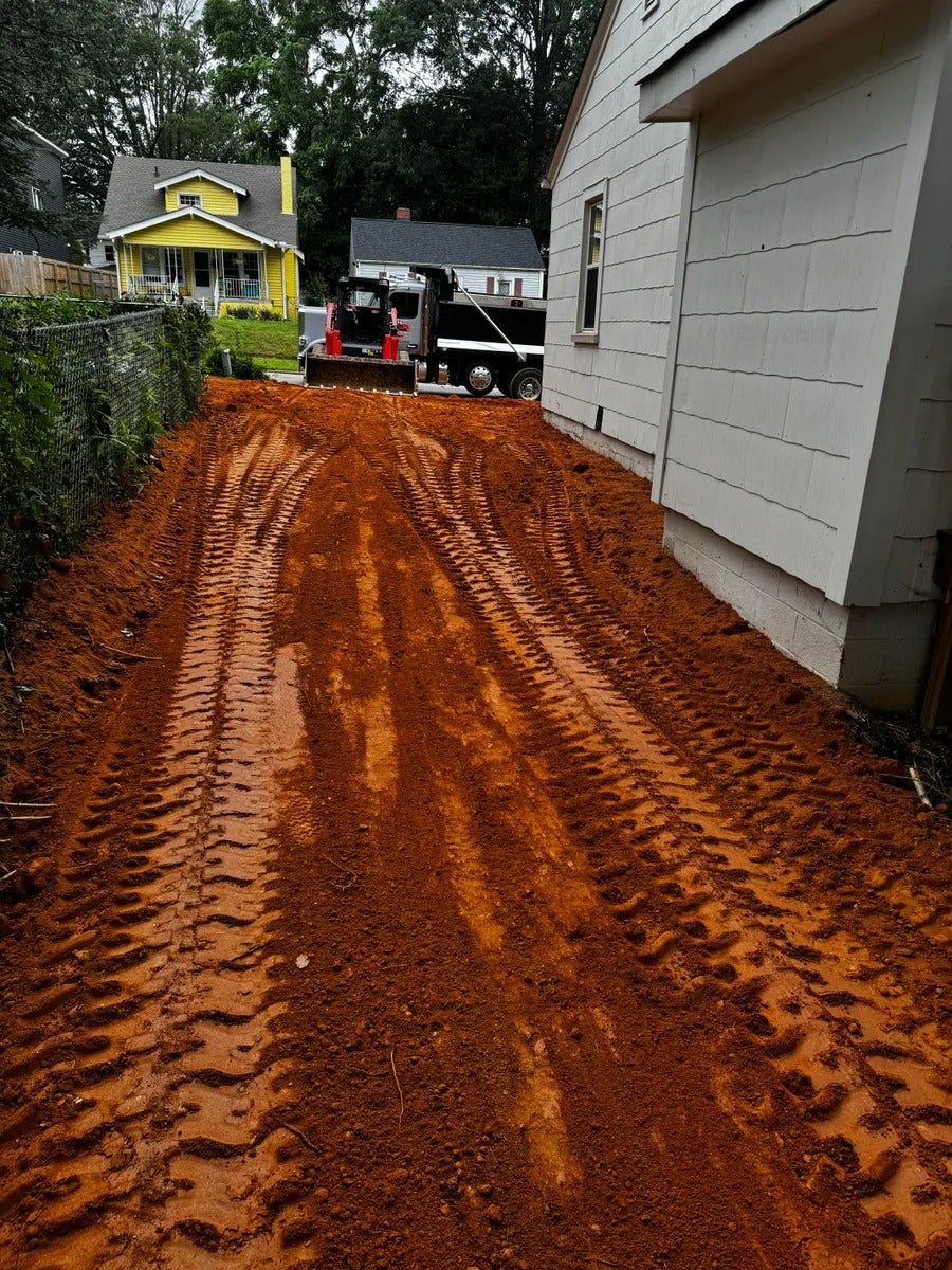 A long, narrow dirt driveway with fresh tire tracks leading to a dump truck and a backhoe between two houses.
