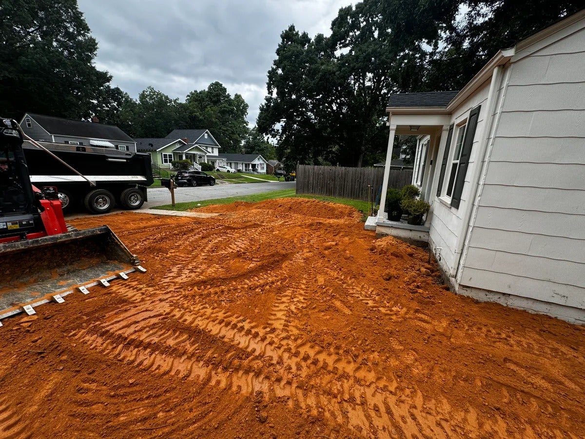 Red dirt laid by a small excavator and dump truck around a white house on an overcast day.