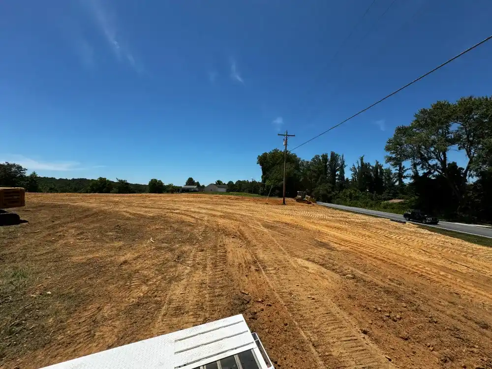 A large, cleared brown dirt lot with tire tracks, houses and trees in the distance, under a clear blue sky.