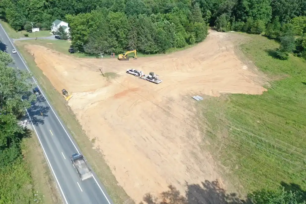 Aerial view of land cleared for construction next to a road, with excavators, a trailer, and concrete pipes.